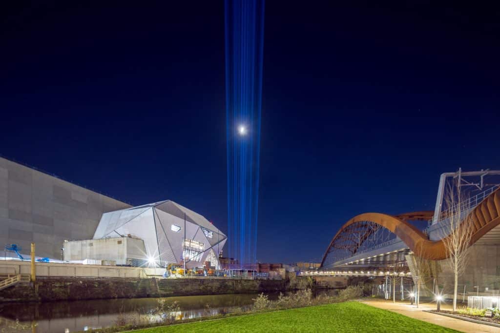 blue-light-beams-shotting-up-towards-sky-between-factory-international-and-bridge