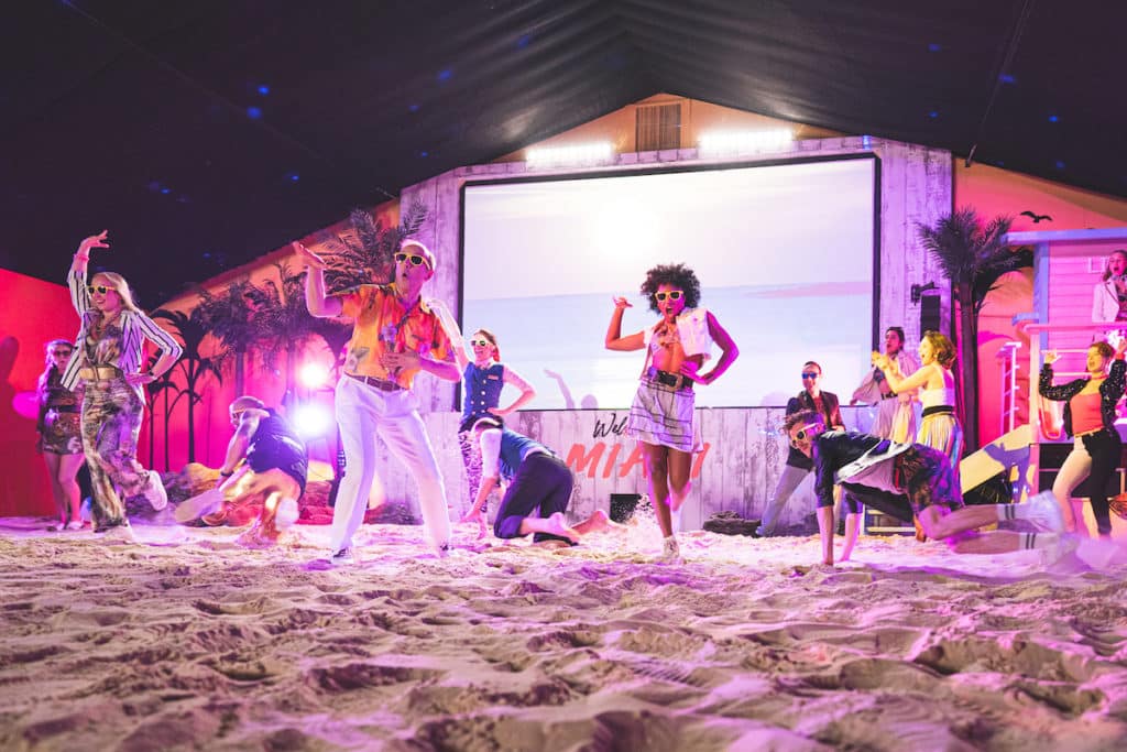 a group in 80's beach wear dances in front of a big screen