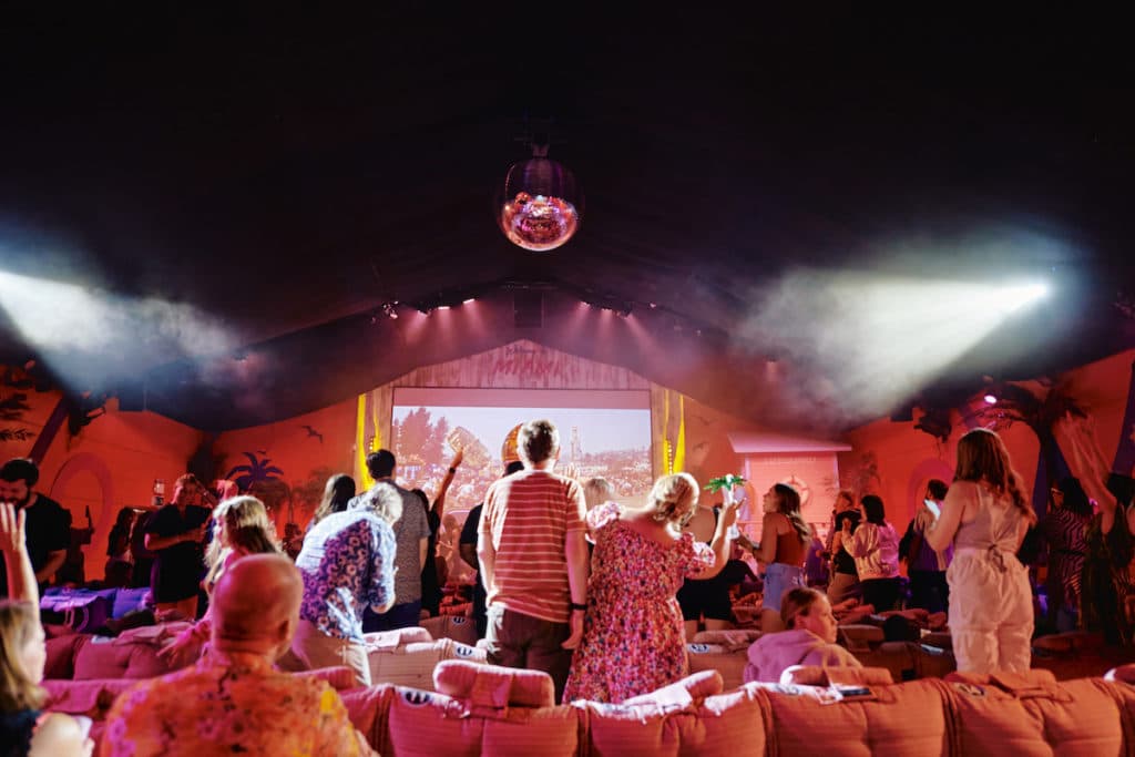 people dance in pink lighting in front of a big screen surrounded by palm trees and with a disco ball above